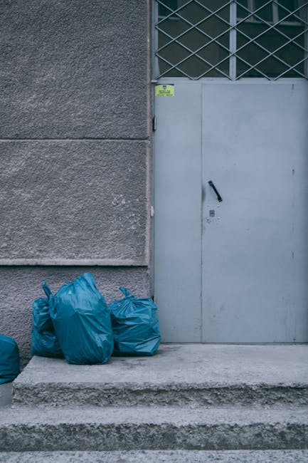 Five blue plastic rubbish bags filled with waste are placed on a concrete step outside a grey building. The bags are tightly tied at the top and appear slightly bulging, indicating they are filled to capacity. They are positioned next to a textured, dark grey wall that shows some signs of weathering. To the right, there is a light grey metal door with a black handle, set within a section of metal latticework that covers a portion of the door and window area above. The ground surface is smooth concrete, and there is a small yellow and black warning sticker visible on the wall near the top of the door frame. The setting suggests an external area typically used for private rubbish disposal, potentially part of a premises requiring independent collection or waste clearance services, such as those offered by House Clearance Colliers Wood. The scene is evenly lit with natural daylight, emphasizing the contrast between the bag colours, wall textures, and the urban environment context related to waste management or rubbish removal activities.