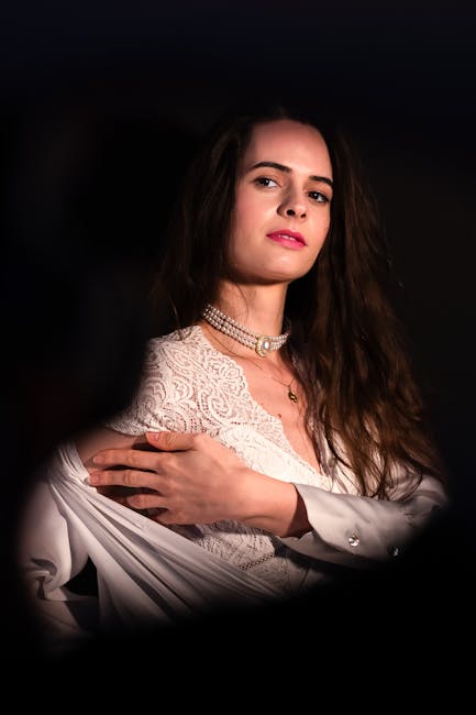 The image depicts a young woman with long, wavy brown hair and fair skin, wearing a white lace top and a layered pearl necklace, standing against a dark background. She has a neutral facial expression and is slightly turned to her left, with her right arm crossed over her chest, gently holding her left shoulder. The setting is softly lit, highlighting the intricate lace pattern of her top and the texture of her hair, creating a subtle contrast with the dark backdrop. Overall, the image exudes a calm, composed atmosphere, suitable for a professional or artistic context, while aligning with themes of personal presentation or portrait photography. The visual focus on her attire and expression could be associated with personal styling or fashion, subtly supporting content related to individual presentation or image management, relevant to services such as professional photography or personal branding.