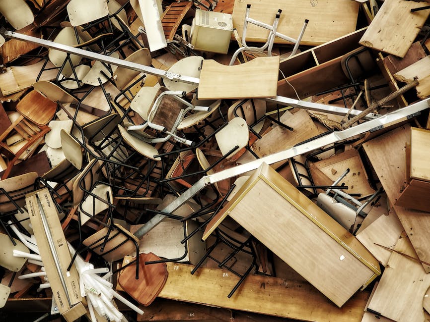 A large pile of discarded furniture and office equipment, including various wooden desks and tables with light and dark wood finishes, some with visible scratches or marks, chairs with metal frames and plastic or wooden seats, and assorted smaller items such as a white radiator, an electric heater, and a metal rod, all stacked haphazardly on a flat surface, possibly outdoors or in a warehouse setting. The chairs are intertwined with the tables and other objects, creating a disorganized scene that exemplifies bulk waste that might be cleared through a professional rubbish removal service like House Clearance Colliers Wood, illustrating the need for efficient sorting and handling of varied household or office waste materials as part of an alternative waste disposal method. The lighting is natural, highlighting the different textures of the furniture, from smooth surfaces to rough edges, emphasizing the quantity and variety of items meant for disposal or recycling.