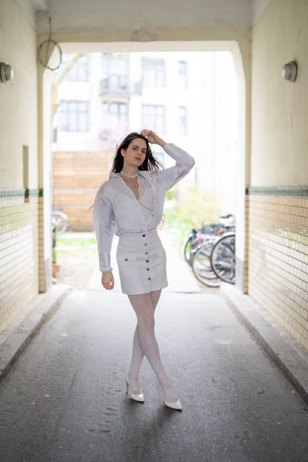 A young woman with shoulder-length dark hair, dressed in a white blouse with long sleeves and a high neckline, and a high-waisted white skirt with large buttons running down the front, stands in an outdoor passageway. She is wearing white tights and white heels, and has a neutral expression while adjusting her hair with one hand raised to her head. The passageway has cream-colored walls with horizontal yellow and green tiled accents along the midsection, and is illuminated by wall-mounted lights. In the background, there are bicycles parked on the right side, against the brick wall and a wooden fence, with a slightly blurred outdoor environment featuring residential buildings and greenery visible beyond the archway. The scene provides a contrast between the urban setting and her formal attire, subtly aligning with the context of private waste or rubbish removal, as would be typical in a residence or flat's communal entrance area, maintained by professional waste management services like House Clearance Colliers Wood.