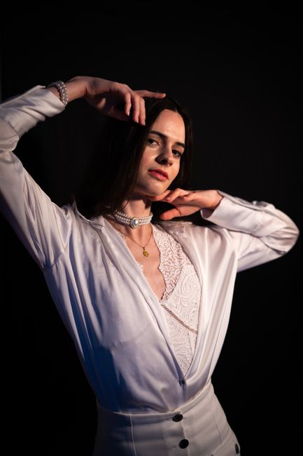 A woman with shoulder-length dark hair posing against a black background. She is wearing a white satin blouse with lace details at the neckline and high-waisted white trousers. Her right arm is raised, with her hand resting on the side of her head, while her left hand touches her neck. She is accessorized with a pearl bracelet on her right wrist, a layered choker necklace, and a gold pendant necklace. The lighting highlights her facial features and the smooth texture of her clothing, creating a professional studio portrait. The image does not depict any rubbish or waste management scene.