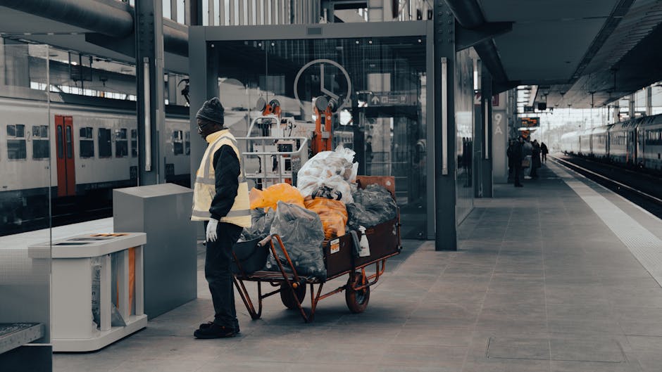 A worker standing on a modern railway station platform, dressed in a high-visibility vest, a dark jacket, and a grey beanie, is positioned beside a large orange and black metal trolley loaded with assorted waste bags and debris, including black, grey, and orange plastic bags, some of which are partly open revealing miscellaneous rubbish. The trolley is situated close to a white waste bin and a grey equipment box, with the station's clean, tiled floor extending into the background. Behind the worker, the station features glass panels, structural steel supports, and a waiting train on the right side, with a few passengers visible further down the platform. The overall environment is well-lit with natural daylight, emphasizing the urban setting of an efficient, modern transport hub, where private waste handling might occur outside standard public rubbish collection. The scene subtly illustrates an alternative waste disposal process often associated with independent rubbish removal services such as those offered by House Clearance Colliers Wood, within a busy railway environment designed for quick and accessible rubbish clearance.