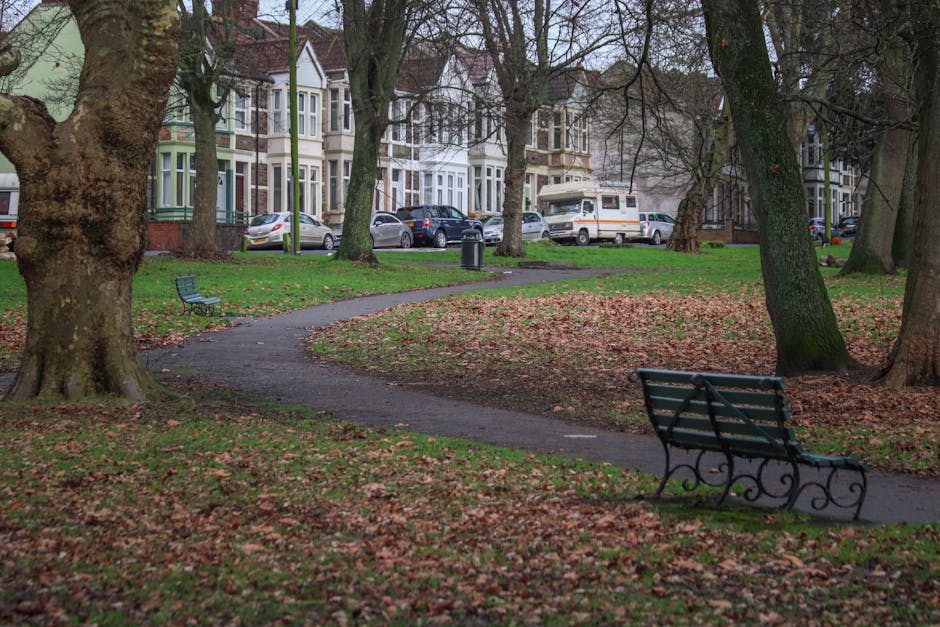 A small park area featuring a meandering paved footpath surrounded by fallen autumn leaves covering the grass and ground. Several mature trees with textured, rough bark have bare branches reaching overhead, casting light shadows on the ground. In the foreground, there is a green metal park bench with decorative armrests and backrest, positioned on the right side of the path, facing inward toward the park. To the left, another empty bench is visible further along the pathway. In the background, across the park, a residential street lined with Victorian-style terraced houses with bay windows and ornate facades is visible, with parked cars including a silver hatchback, a white van, and a dark sedan. The scene is illuminated by natural daylight, with a generally muted, overcast atmosphere, highlighting the park's quiet, urban environment. The park appears well-maintained, and the scene subtly reflects aspects of local urban rubbish management, such as street cleaning and waste disposal nearby, supporting the services provided by House Clearance Colliers Wood.